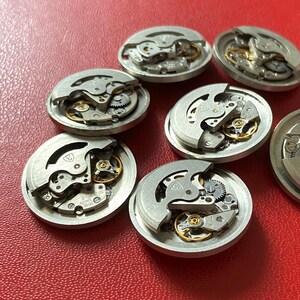 Puede incluir: Una colecci&oacute;n de movimientos de reloj desmontados, mostrando intrincados engranajes, muelles y volantes. Los componentes plateados est&aacute;n dispuestos sobre una superficie roja, destacando la ingenier&iacute;a de precisi&oacute;n de los relojes.