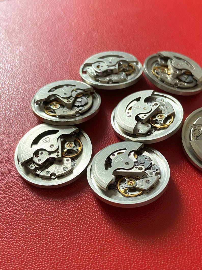 Puede incluir: Una colecci&oacute;n de movimientos de reloj desmontados, mostrando intrincados engranajes, muelles y volantes. Los componentes plateados est&aacute;n dispuestos sobre una superficie roja, destacando la ingenier&iacute;a de precisi&oacute;n de los relojes.