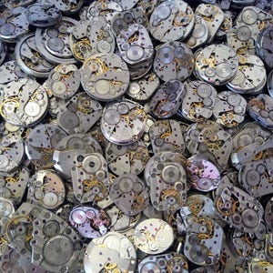 May include: A close-up image of a pile of silver watch movements. The watch movements are all different sizes and shapes, and they are all made of metal. The watch movements are all very intricate and detailed.