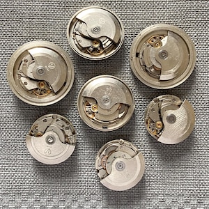 May include: Seven disassembled watch movements are displayed, revealing intricate mechanical parts. The silver-coloured metal components include gears, springs, and balance wheels. The watch movements are arranged on a textured grey surface.