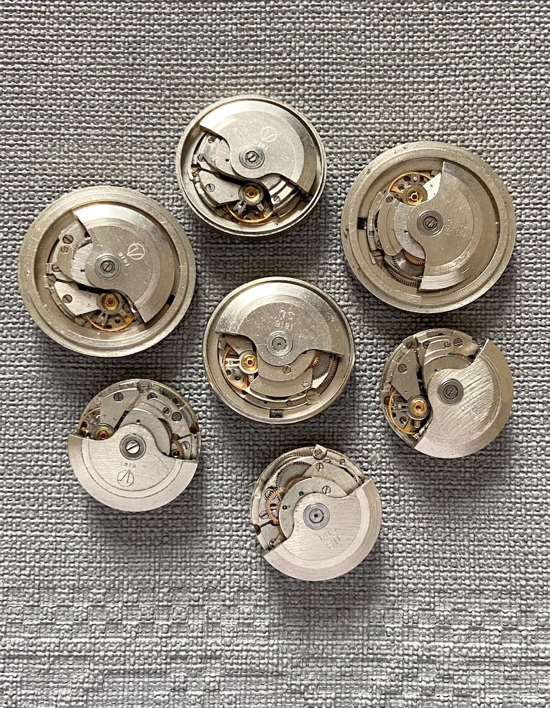 May include: Seven disassembled watch movements are displayed, revealing intricate mechanical parts. The silver-coloured metal components include gears, springs, and balance wheels. The watch movements are arranged on a textured grey surface.