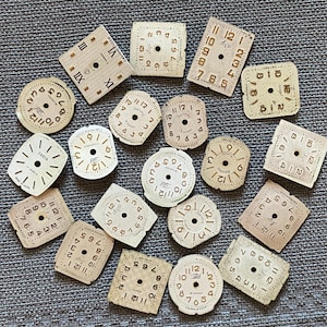 May include: A collection of vintage watch faces in various shapes, including round and square. The faces are off-white with black numerals and markings. Some faces have text, including "Zaria" and "17 Jewels".