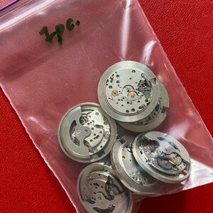 Puede incluir: Una bolsa de pl&aacute;stico transparente que contiene varios movimientos de reloj plateados. Los movimientos son redondos con intrincados engranajes y componentes internos. La bolsa est&aacute; sobre una superficie roja y el texto "3pc." es visible.