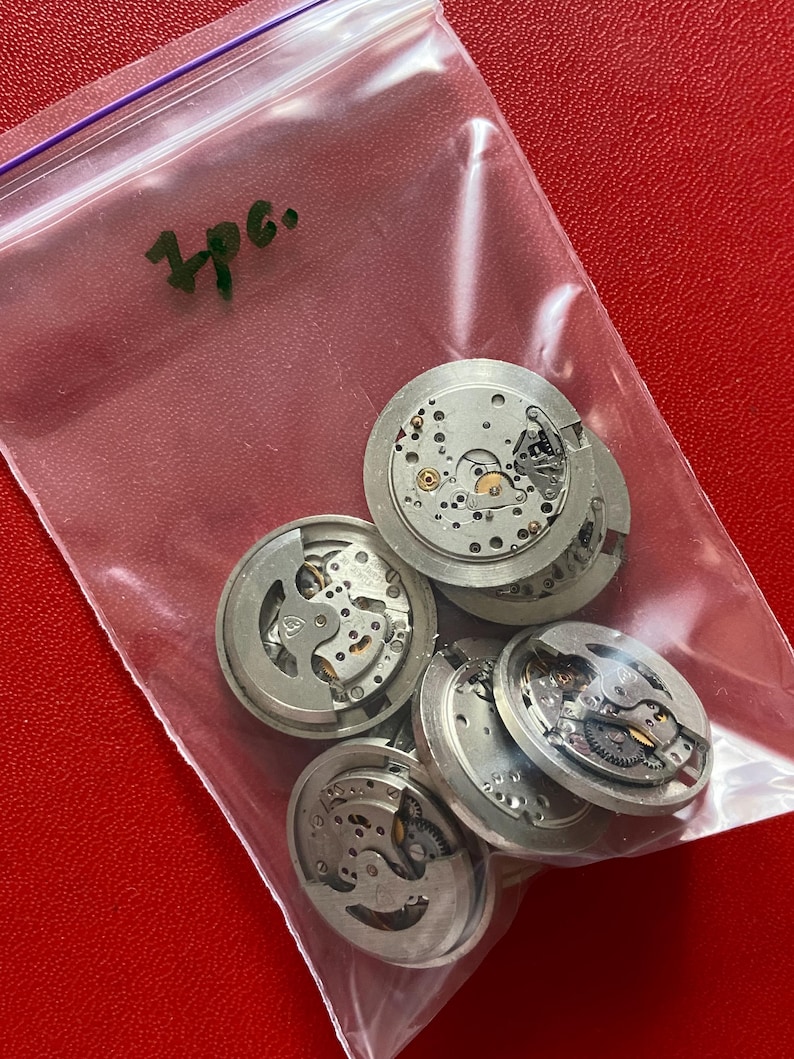 Puede incluir: Una bolsa de pl&aacute;stico transparente que contiene varios movimientos de reloj plateados. Los movimientos son redondos con intrincados engranajes y componentes internos. La bolsa est&aacute; sobre una superficie roja y el texto "3pc." es visible.