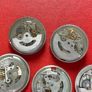 Puede incluir: Una colecci&oacute;n de movimientos de reloj desmontados, que muestra intrincados engranajes, resortes y volantes. Los componentes plateados est&aacute;n dispuestos sobre un fondo rojo texturizado, destacando los detalles mec&aacute;nicos de cada mecanismo de reloj.