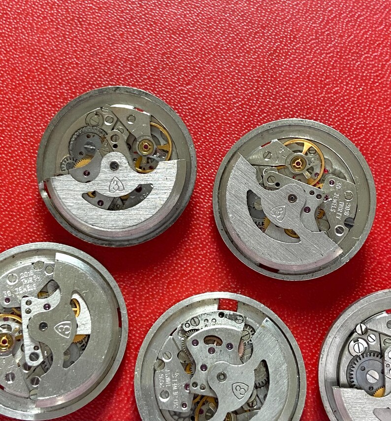 Puede incluir: Una colecci&oacute;n de movimientos de reloj desmontados, que muestra intrincados engranajes, resortes y volantes. Los componentes plateados est&aacute;n dispuestos sobre un fondo rojo texturizado, destacando los detalles mec&aacute;nicos de cada mecanismo de reloj.