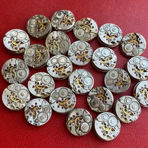 Peut inclure: Une collection de 20 mouvements de montres argent&eacute;s avec des engrenages complexes et des rubis. Les mouvements de montres sont dispos&eacute;s en cercle sur une surface rouge.