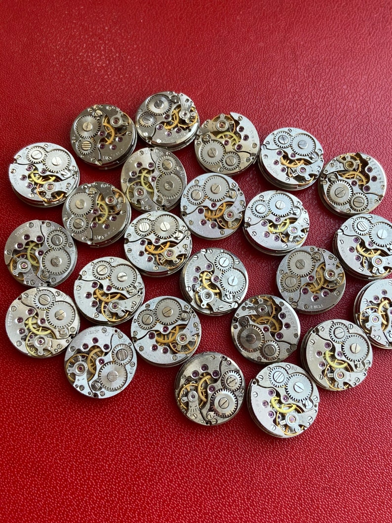 Peut inclure: Une collection de 20 mouvements de montres argent&eacute;s avec des engrenages complexes et des rubis. Les mouvements de montres sont dispos&eacute;s en cercle sur une surface rouge.