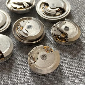 May include: A selection of dismantled watch movements, displaying complex internal mechanisms. The silver components, including gears, springs, and balance wheels, reveal the inner workings of timepieces. The parts are arranged on a textured, grey surface.