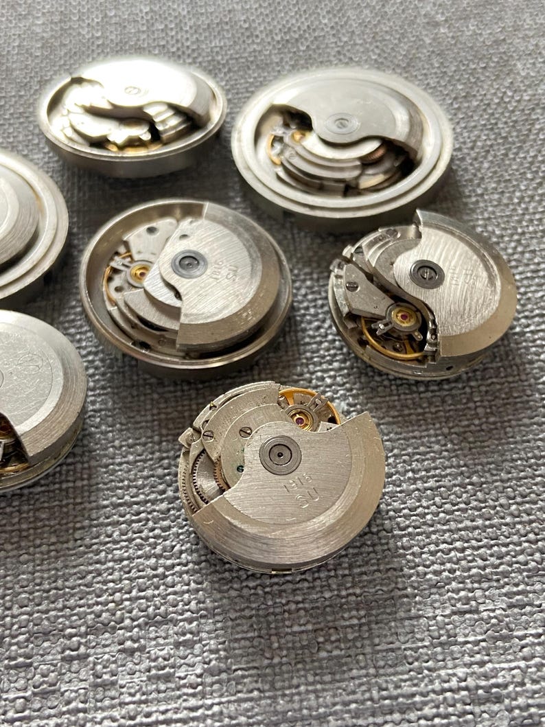 May include: A selection of dismantled watch movements, displaying complex internal mechanisms. The silver components, including gears, springs, and balance wheels, reveal the inner workings of timepieces. The parts are arranged on a textured, grey surface.