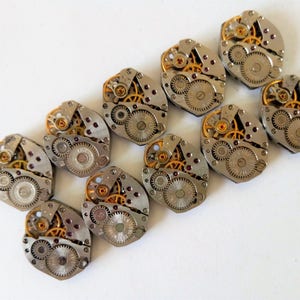 May include: Ten silver and gold watch movements with intricate gears and red jewels. The watch movements are arranged in a staggered pattern on a white surface.