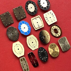 May include: A collection of vintage watch faces in various shapes and colors, including black, white, gold, and blue. Some faces feature Roman numerals, while others have a more minimalist design. The watch faces are arranged on a red surface.