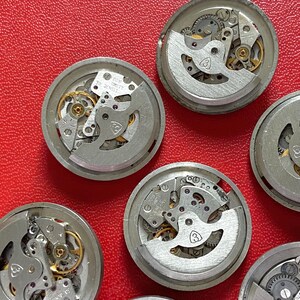 Puede incluir: Primer plano de varios movimientos de reloj desmontados, con engranajes, muelles y otros componentes internos visibles. Las piezas plateadas presentan detalles dorados. Los movimientos est&aacute;n dispuestos sobre una superficie roja. Algunos tienen la inscripci&oacute;n "20 Jewels".