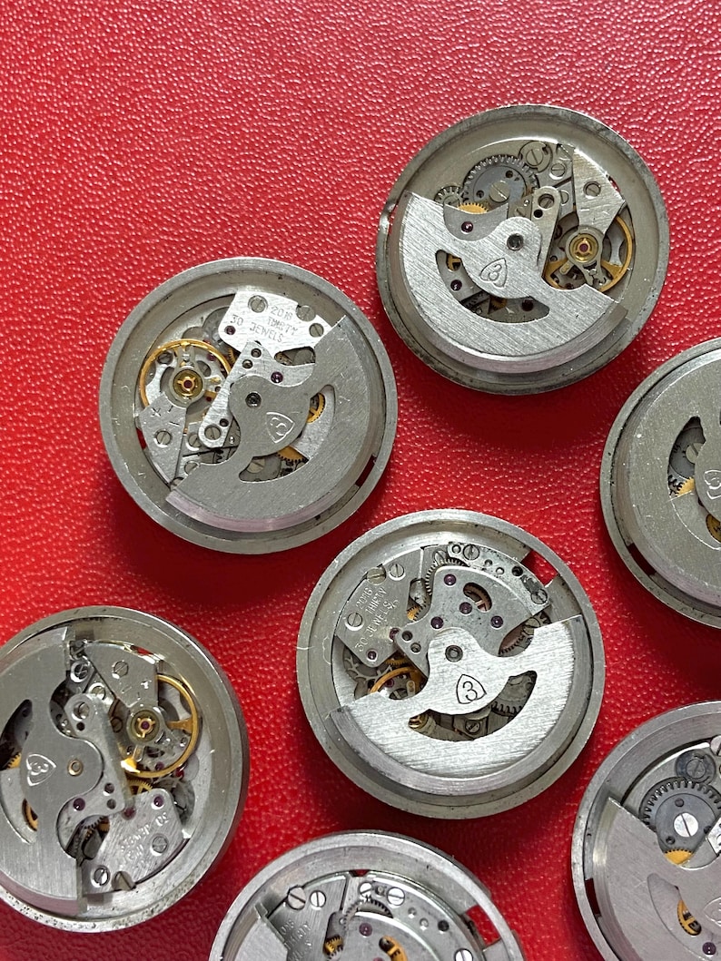 Puede incluir: Primer plano de varios movimientos de reloj desmontados, con engranajes, muelles y otros componentes internos visibles. Las piezas plateadas presentan detalles dorados. Los movimientos est&aacute;n dispuestos sobre una superficie roja. Algunos tienen la inscripci&oacute;n "20 Jewels".
