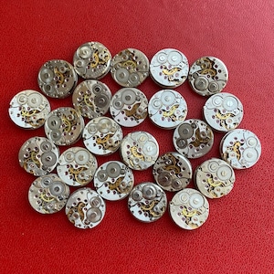 Peut inclure: Une collection de 25 mouvements de montres argent&eacute;s avec des engrenages et des ressorts complexes. Les mouvements de montres sont dispos&eacute;s en cercle sur une surface rouge.