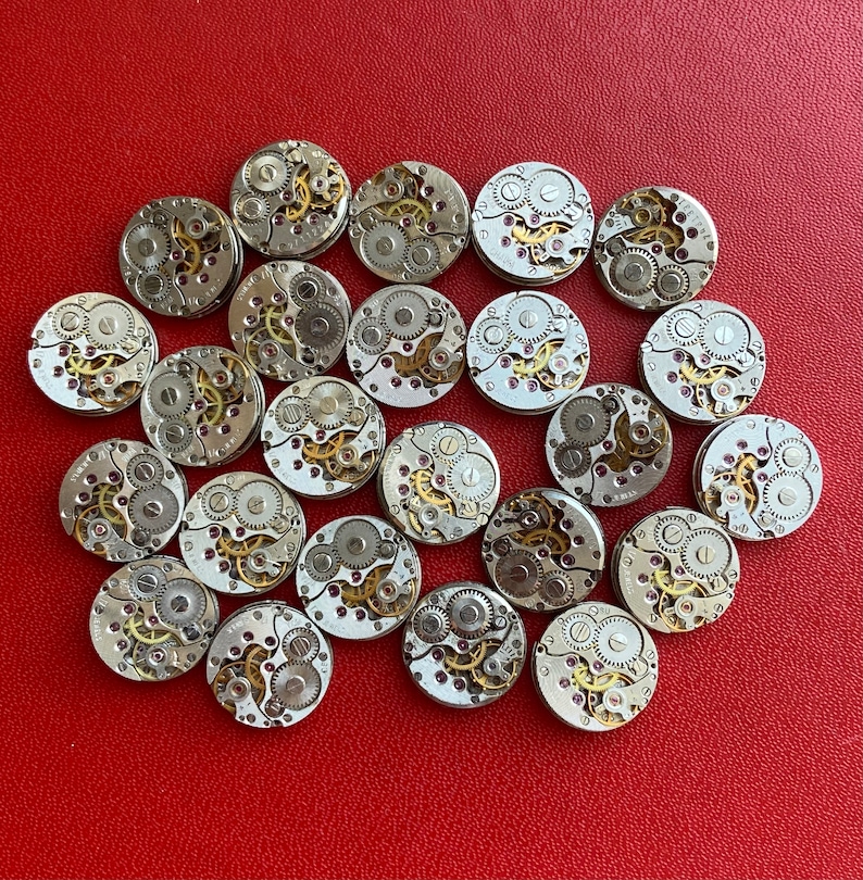 Peut inclure: Une collection de 25 mouvements de montres argent&eacute;s avec des engrenages et des ressorts complexes. Les mouvements de montres sont dispos&eacute;s en cercle sur une surface rouge.
