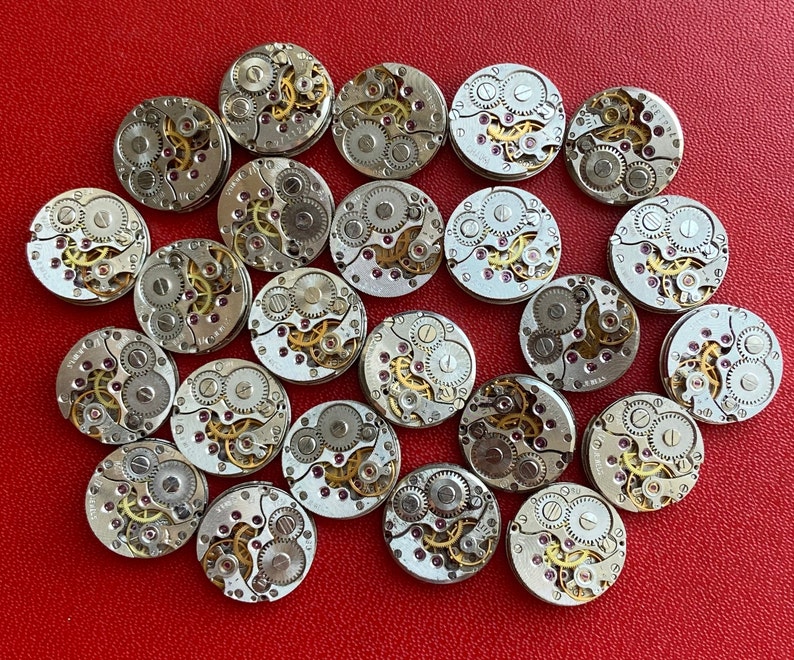 Peut inclure: Une collection de 20 mouvements de montres argent&eacute;s, chacun avec des engrenages complexes et des motifs complexes. Les mouvements de montres sont dispos&eacute;s en cercle sur une surface rouge.