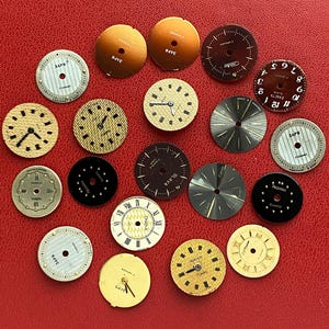 Peut inclure: Une collection de cadrans de montres vintage de différentes couleurs et designs, dont l'or, l'argent et le marron. Certains cadrans présentent des chiffres romains, tandis que d'autres ont des chiffres arabes ou des marqueurs simples. Les cadrans sont disposés sur une surface rouge.