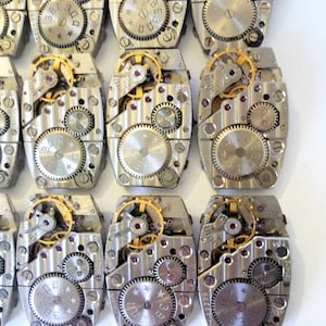 May include: A collection of 10 silver-toned watch movements with intricate gears and a star-shaped design. The movements are arranged in two rows of five, with the top row slightly offset from the bottom row.