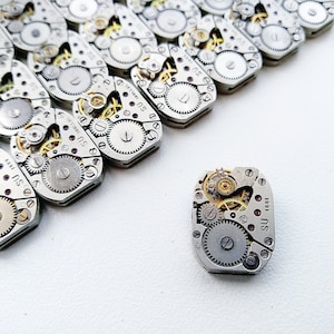 May include: A collection of vintage watch movements, displaying intricate internal mechanisms. The silver-coloured metal components, including gears and springs, are arranged on a white surface, demonstrating the detailed craftsmanship of watchmaking.