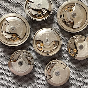 May include: Seven silver watch movements displaying intricate internal mechanisms with visible gears and springs. The watch parts are arranged on a textured grey surface, revealing the detailed craftsmanship of each component.