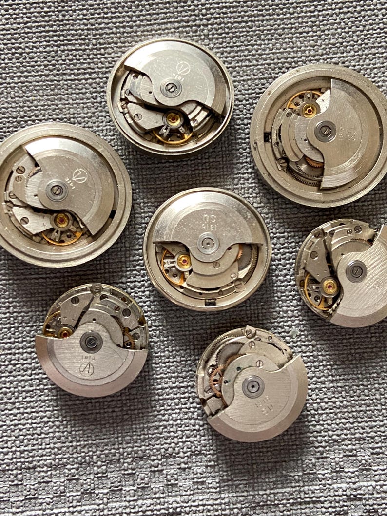 May include: Seven silver watch movements displaying intricate internal mechanisms with visible gears and springs. The watch parts are arranged on a textured grey surface, revealing the detailed craftsmanship of each component.