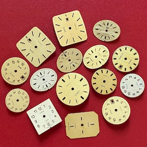 May include: A collection of vintage watch faces in various shapes and sizes, including round, oval, and rectangular. The faces are primarily gold-toned with black numerals and markings. Some faces display the brand name "ZARIA" or "34PB". The background is a solid red.
