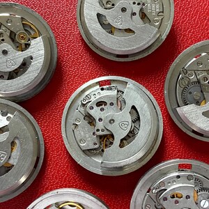 Puede incluir: Primer plano de varios movimientos de reloj desmontados, mostrando intrincados engranajes, muelles y joyas. Los componentes plateados est&aacute;n dispuestos sobre una superficie roja, resaltando los detalles mec&aacute;nicos de los mecanismos del reloj. Algunos movimientos tienen el texto "30 JEWELS".