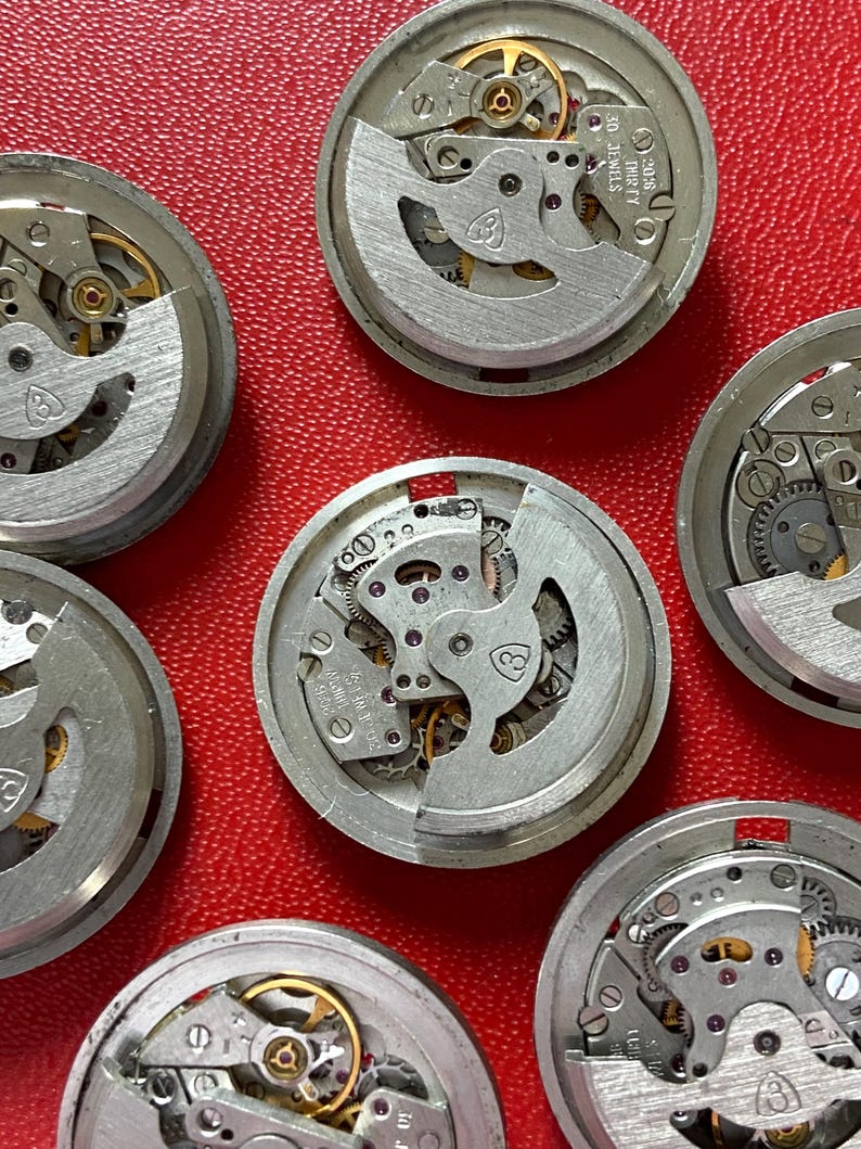 Puede incluir: Primer plano de varios movimientos de reloj desmontados, mostrando intrincados engranajes, muelles y joyas. Los componentes plateados est&aacute;n dispuestos sobre una superficie roja, resaltando los detalles mec&aacute;nicos de los mecanismos del reloj. Algunos movimientos tienen el texto "30 JEWELS".