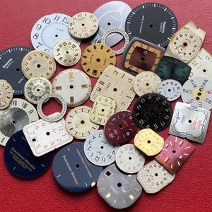 Puede incluir: Una colección de esferas de reloj antiguas en diversas formas y colores, incluyendo blanco, negro, dorado y burdeos. Las esferas muestran números, índices y nombres de marcas. Algunas son redondas, cuadradas o rectangulares, con signos de desgaste.