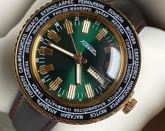 May include: A gold-tone wristwatch with a green face and black leather band. The watch face features a world time dial with city names in Cyrillic script and a date window. The watch is in a box.