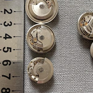 May include: Close-up of several disassembled watch movements, showcasing intricate mechanical components. The silver-toned parts include gears, springs, and other small elements. A ruler is placed next to the watch parts for scale.
