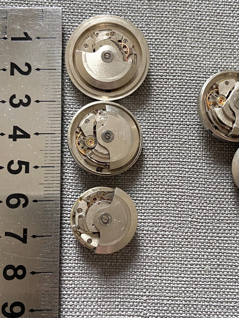 May include: Close-up of several disassembled watch movements, showcasing intricate mechanical components. The silver-toned parts include gears, springs, and other small elements. A ruler is placed next to the watch parts for scale.