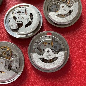 Puede incluir: Una colecci&oacute;n de movimientos de reloj desmontados, que muestra intrincados componentes mec&aacute;nicos. Las piezas met&aacute;licas plateadas, incluyendo engranajes y muelles, est&aacute;n dispuestas sobre una superficie roja. Los movimientos del reloj son circulares y tienen un dise&ntilde;o parcialmente expuesto.