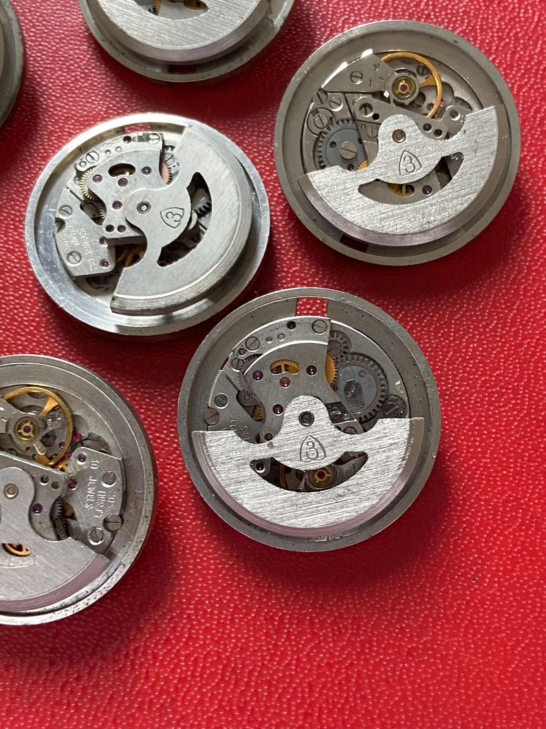 Puede incluir: Una colecci&oacute;n de movimientos de reloj desmontados, que muestra intrincados componentes mec&aacute;nicos. Las piezas met&aacute;licas plateadas, incluyendo engranajes y muelles, est&aacute;n dispuestas sobre una superficie roja. Los movimientos del reloj son circulares y tienen un dise&ntilde;o parcialmente expuesto.