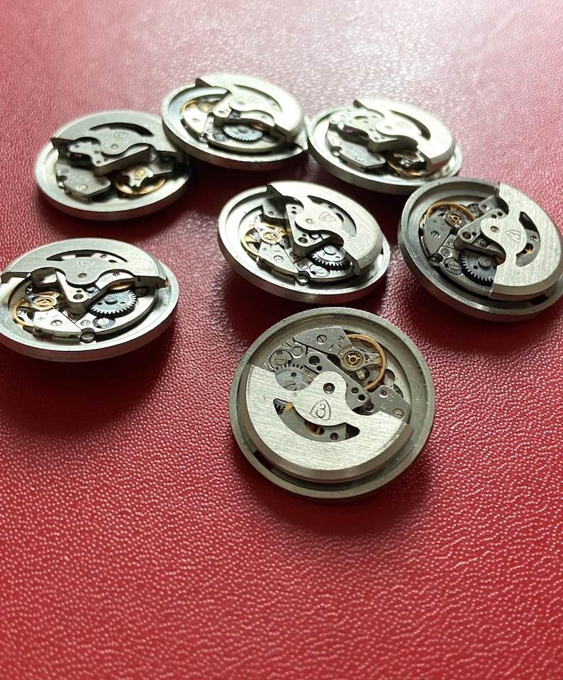 Puede incluir: Varios movimientos de reloj desmontados, mostrando engranajes, muelles y otros componentes internos. Las piezas met&aacute;licas plateadas est&aacute;n dispuestas sobre una superficie roja, revelando la compleja mec&aacute;nica de los dispositivos de medici&oacute;n del tiempo.