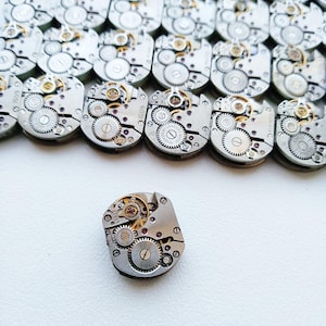 May include: A close-up of a group of silver watch movements with intricate gears and a single movement in the foreground.