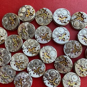 Peut inclure: Une collection de 20 mouvements de montres argent&eacute;s avec des engrenages complexes et des rubis. Les mouvements de montres sont dispos&eacute;s en cercle sur une surface rouge.