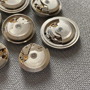 May include: Several silver watch movements with exposed gears and internal mechanisms. The watch components are displayed on a textured grey surface, highlighting the mechanical details. Some have the text "1616 SU" on them.