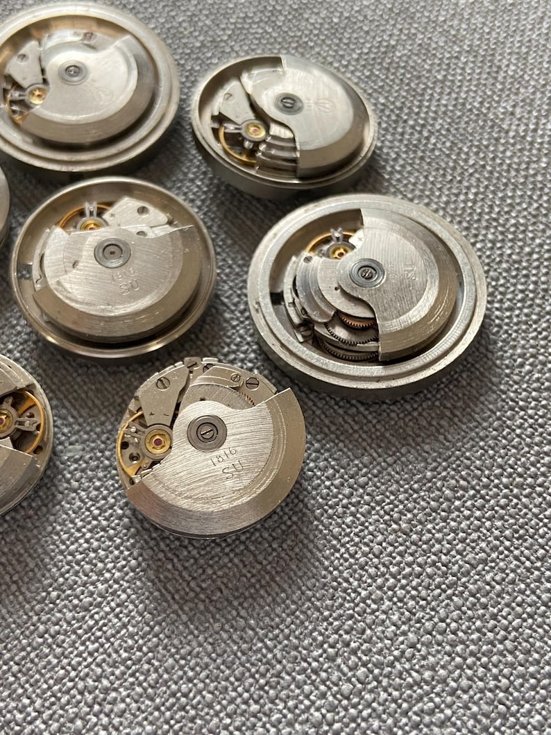 May include: Several silver watch movements with exposed gears and internal mechanisms. The watch components are displayed on a textured grey surface, highlighting the mechanical details. Some have the text "1616 SU" on them.