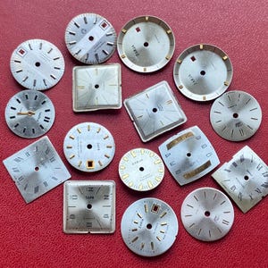 Könnte beinhalten: Eine Sammlung von Vintage-Zifferblättern in verschiedenen Formen, darunter rund, quadratisch und rechteckig. Die Zifferblätter sind silberfarben mit goldenen Akzenten und schwarzen Ziffern. Einige Zifferblätter tragen die Marke "Zaria" oder "Luch". Der Hintergrund ist einfarbig rot.