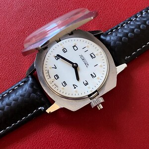 May include: A silver-toned wristwatch with a white dial, black numerals, and black hands. The watch features a hinged metal cover over the crystal and a black textured leather strap with white stitching. The dial displays the text Raketa and CCCP.