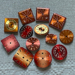 Puede incluir: Una colección de doce esferas de reloj vintage de diversas formas, incluyendo cuadradas, redondas y rectangulares. Las esferas presentan acabados metálicos en tonos cobre, bronce y rojo, con números impresos, índices y el texto Zaria y CCCP.