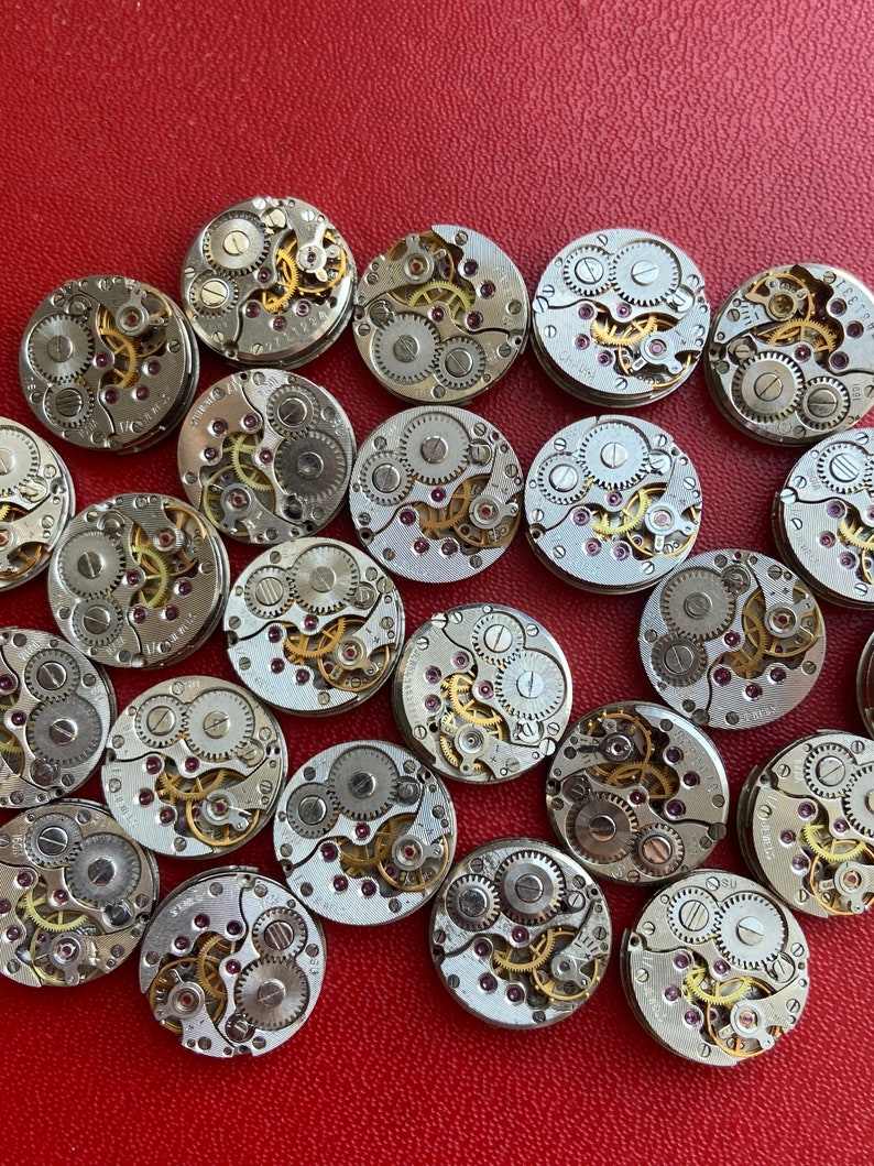 Peut inclure: Une collection de 20 mouvements de montres argent&eacute;s avec des engrenages complexes et des rubis. Les mouvements de montres sont dispos&eacute;s en cercle sur une surface rouge.