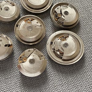 May include: A selection of dismantled watch movements, displaying detailed gears and mechanical elements. The silver-coloured metal components are arranged on a textured grey surface, revealing the inner workings of timepieces.