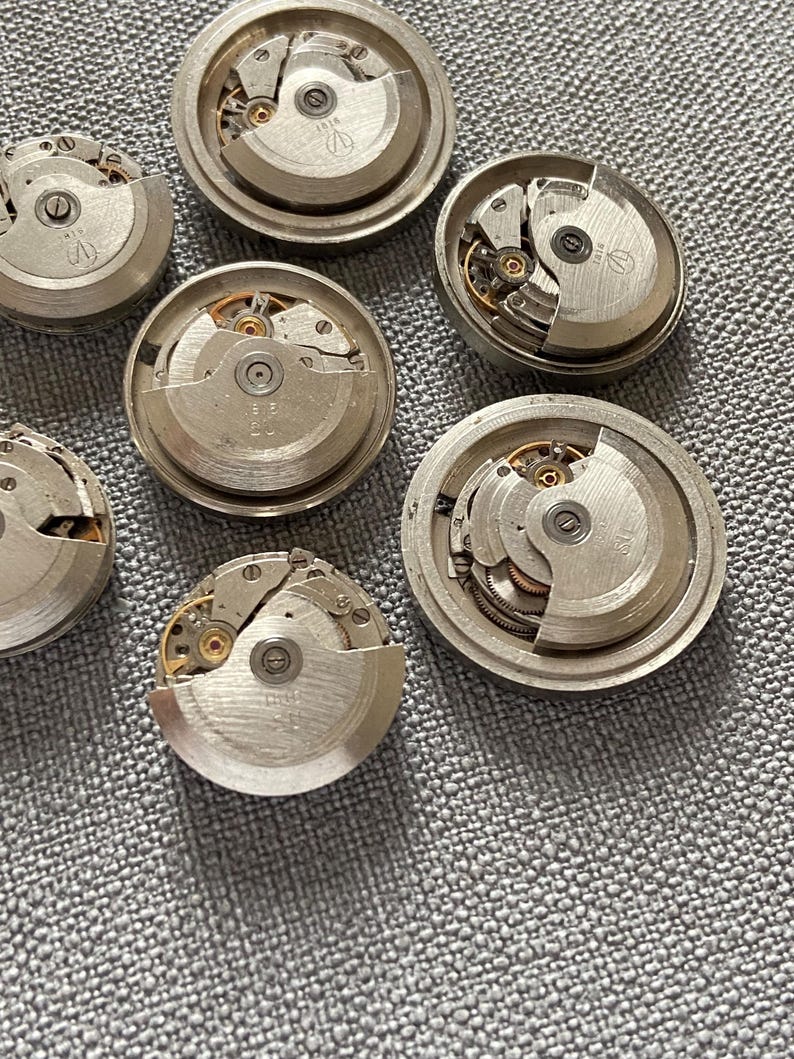 May include: A selection of dismantled watch movements, displaying detailed gears and mechanical elements. The silver-coloured metal components are arranged on a textured grey surface, revealing the inner workings of timepieces.