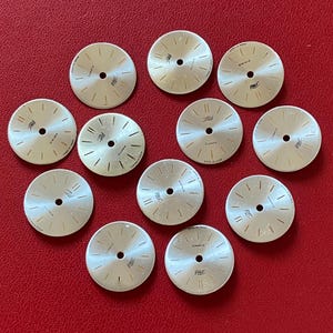 Puede incluir: Una colección de once esferas de reloj circulares de tono plateado, cada una con un orificio central, marcadores de hora negros finos y el texto '27 Jewels' y 'MADE IN USSR' impreso en la superficie. Las esferas están sobre una superficie roja.