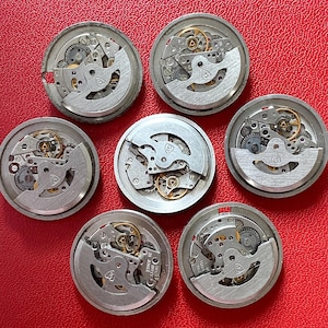Puede incluir: Colecci&oacute;n de siete mecanismos de reloj desmontados, cada uno con intrincados engranajes, resortes y otros componentes internos. Las piezas del reloj est&aacute;n dispuestas sobre una superficie roja.