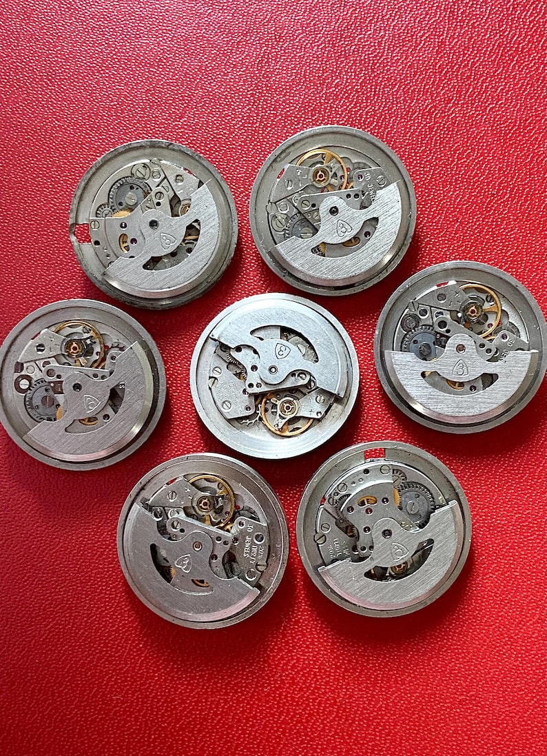 Puede incluir: Colecci&oacute;n de siete mecanismos de reloj desmontados, cada uno con intrincados engranajes, resortes y otros componentes internos. Las piezas del reloj est&aacute;n dispuestas sobre una superficie roja.