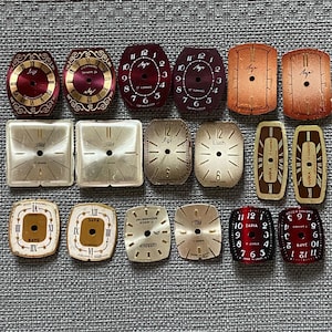 Könnte beinhalten: Eine Sammlung von achtzehn Vintage-Uhrenzifferblättern in verschiedenen Formen, Farben und Designs. Die Zifferblätter weisen unterschiedliche Ziffernstile auf, darunter römische und arabische Ziffern, mit Markennamen wie Luch, Zaria und Cornavin.